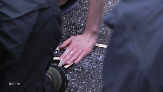 Blick zwischen zwei Schulter von Personen mit Uniform hindurch auf den Straßenasphalt auf dem eine Hand einer weiteren person festgeklebt ist. © Screenshot 