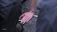 Blick zwischen zwei Schulter von Personen mit Uniform hindurch auf den Straßenasphalt auf dem eine Hand einer weiteren person festgeklebt ist. © Screenshot 