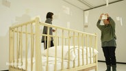 In einem weißgestrichenen Ausstellungsraum steht ein Hochstallbett für Säuglinge, im Hintergrund hängen zwei Frauen Zettel an von der Decke hängenede Fäden auf. © Screenshot 