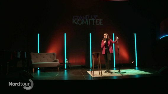 Auf einer kleineren Bühne steht eine Frau mit Mikrofon im Scheinwerferlicht. © Screenshot 