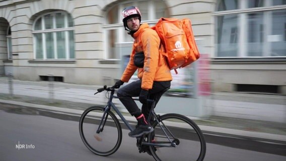 Ein Fahrradlieferand blickt sich während der Fahrt nach hinten um. Er tägt eine prominente orange-farbene Jacke und einen gleichfarbigen Rucksack. © Screenshot 