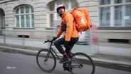 Ein Fahrradlieferand blickt sich während der Fahrt nach hinten um. Er tägt eine prominente orange-farbene Jacke und einen gleichfarbigen Rucksack. © Screenshot 