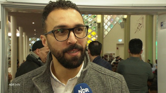 Ein Mann im Interview, im Hintergrund andere in den Innenräumen einer Moschee. © Screenshot 