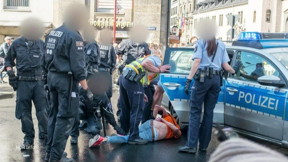 Mehrere Einsatzkräfte der Polizei stehen um einen auf dem Straßenasphalt liegenden Mann mit auf den Rücken gefesselten Händen vor einem Einsatzfahrzeug der Polizei herum. © Screenshot 