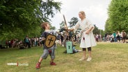 EIn kleinerer Junge und ein Mädchen kämpfen mit Schild und Holzschwertern auf einem Mittelalterfest in einem Park. © Screenshot 