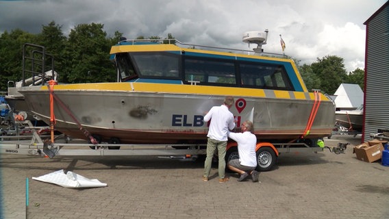 Zwei Männer folieren den Schriftzug "ELBTAXI" auf die Seite eines Motorbootes. © Screenshot 