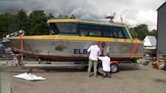 Zwei Männer folieren den Schriftzug "ELBTAXI" auf die Seite eines Motorbootes. © Screenshot 