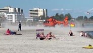 Ein Rettungshubschrauber landet an einem Badestrand. Dabei wirbelt er sehr viel Sand auf. © Screenshot 