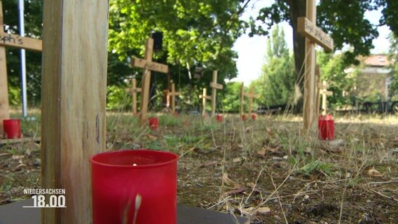 Nachaufnahme einer Grabkerze, im Hintergrund Holzkreuze mit weiteren roten Grabkerzen davor. © Screenshot 