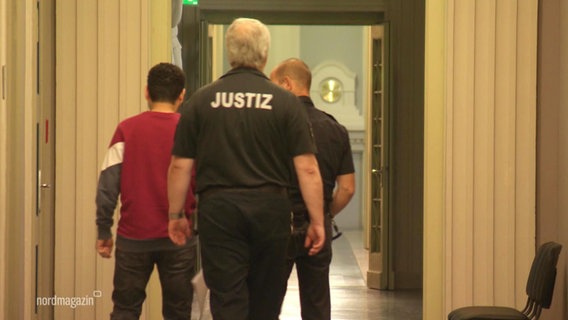 Ein Mann in rotem Pullover wird von zwei Justizbeamten abgeführt. Die eprsonen gehen von der Kamera weg. © Screenshot 