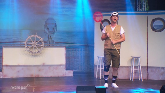 Ein als Angler verkleideter Schauspieler singt auf einer Bühne mit einem Kescher in der Hand tanzend. © Screenshot 
