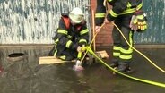 Ein Feuerwehrmann kniet, ein anderer steht auf einer Holzplanke inmitten einer großen Pfütze - sie pumpen mit einer Tauchpumpe das Wasser ab. © Screenshot 