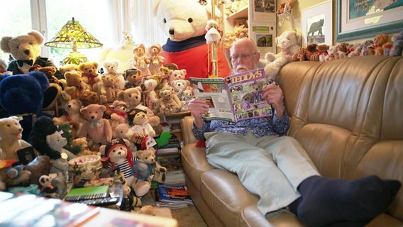 Hans-Dieter Gau sitzt mit ausgestreckten Beinen auf seiner Couch und liest eine Zeitschrift. Um ihn herum sitzen und liegen zahlreiche Teddy-Bären. © Screenshot 