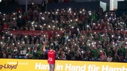 Ein Spieler des TuS Finkenwerder steht vor den Zuschauerrängen und macht eine Bewegung mit dem linken Arm, die Zuschauerinnen und Zuschauer leuchten mit Taschenlampen. © Screenshot 