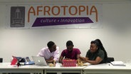 Drei junge Unternehmer:innen unterhalten sich. Hinter ihnen hängt ein Banner des Kulturzentrum "Afrotopia". © Screenshot 