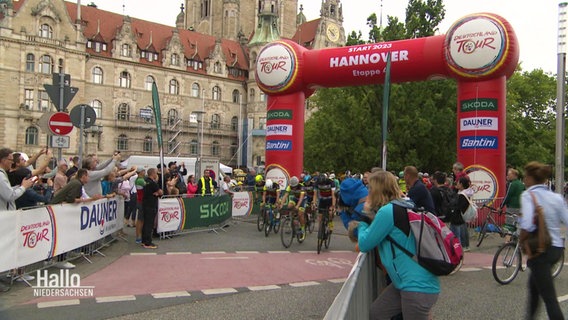 Blick auf den Start von der 4. Etappe der Deutschland Tour 2023 in Hannover: Radsportler fahren durch ein rotes Tor, das den Startpunkt der Etappe markiert. Am Rand der Rennstrecke stehen Zuschauende. © Screenshot 