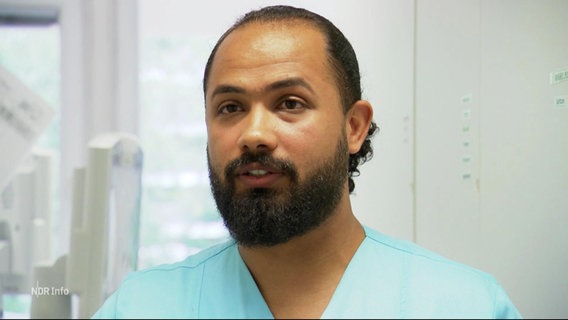 Doktor Abdullah Abdulrahman im Portrait in einem blauen Kasack mit langen dunklen Harren und einem kurzen dunklen Bart. © Screenshot 