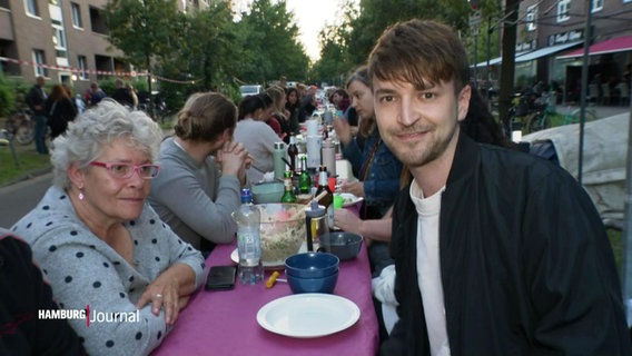 Reporter Tristan Dueck (rechts) sitzt mit unterschiedlichen Menschen an einem gedeckten Tisch mitten auf der Straße. © Screenshot 