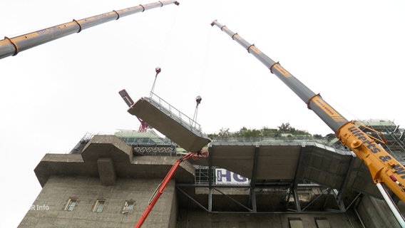 Ein Treppenstück wird mit Hilfe von zwei Kränen auf das Dach eines Bunkers befördert. © Screenshot 