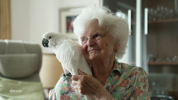 Eine alte Frau streichelt einen weißen Papagei, der auf ihrer rechten Schulter sitzt. © Screenshot 