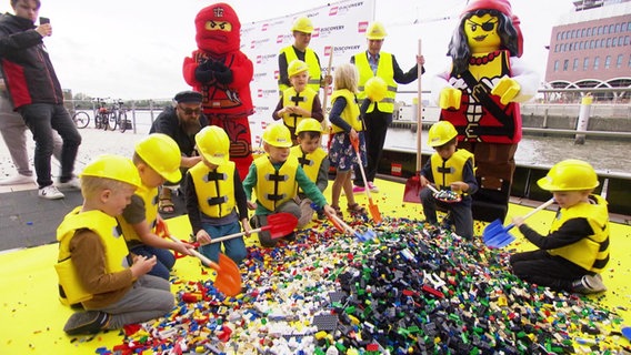 Kita-Kinder in gelben Westen und Bauhelmen stürzen sich mit Schaufeln auf einen großen Berg Lego. © Screenshot 