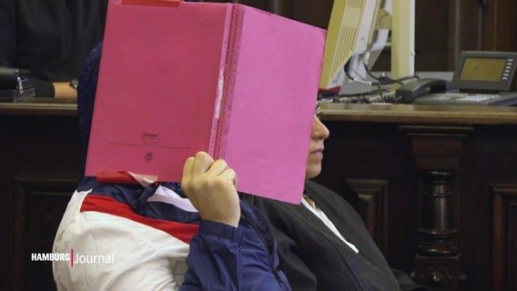 Ein vor Gericht Angeklagter hält sich eine rote Aktenmappe vor das Gesicht. © Screenshot 