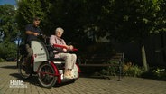 Eine ältere Frau sitzt in einer Fahrrad-Rikscha, die von einem Mann gefahren wird. © Screenshot 