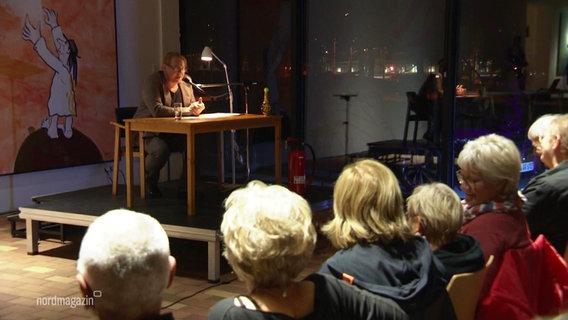 Ben Becker sitzt auf einer Bühne und liest aus einem Buch von Udo Lindenberg. © Screenshot 