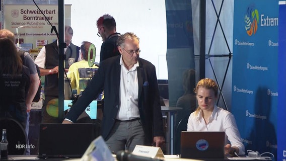 Zwei Personen unterhalten sich über den Schreibtisch gebeugt auf dem Extremwetterkongress in Hamburg. © Screenshot 