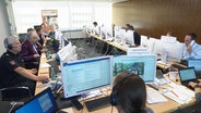 In einem Sitzungsraum sitzen mehrere Menschen, zum Teil in Polizei- oder Bundeswehruniformen, vor Computerbildschirmen. © Screenshot 