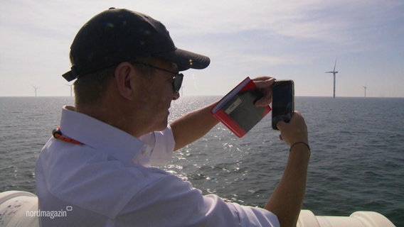 Ein Mann fotografiert mit seinem Handy ein Windrad auf dem Meer. © Screenshot 