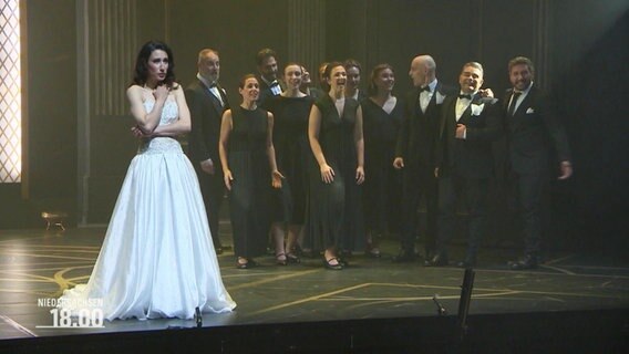 Frau in weißem Kleid auf einer Bühne, im Hintergrund ein Chor. © Screenshot 