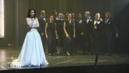 Frau in weißem Kleid auf einer Bühne, im Hintergrund ein Chor. © Screenshot 