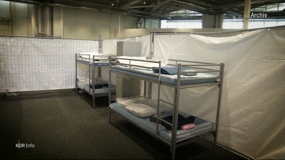 Zwei Stockbetten stehen in einem provisorisch abgehängten Raum in einer Halle. © Screenshot 