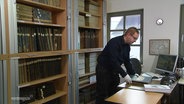 David Krüger steht neben einem Bücherregal und blättert in alten Dokumenten. © Screenshot 