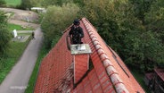 Ein Schornsteinfeger reinigt auf einem Dach stehend einen Schornstein. © Screenshot 