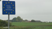 Das Schild "Bundesrepublik Deutschland" mit weißer Schrift auf blauem Grund und den Europa-Sternen darauf an einer Straße, im Hintergrund Felder. © Screenshot 