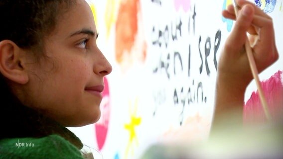 Ein Mädchen steht mit einem Pinsel in der Hand vor einem Wandbild. © Screenshot 
