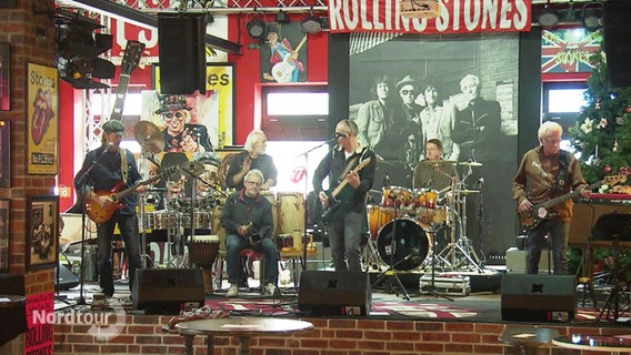 Ältere Herren stehen mit verschiedenen Instrumenten als Rock-Band bei einem Auftritt auf einer Bühne. © Screenshot 