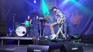 Ein Schlagzeuger und ein Keyboarder performen auf einer Open-Air-Bühne in buntem Scheinwerferlicht. © Screenshot 