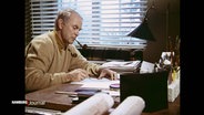 Vicco von Bülow alias Loriot bei der Arbeit: Mit einer Pfeife im Mund sitzt er am Schreibtisch und zeichnet. © Screenshot 