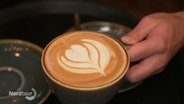 In einer Cappuccino-Tasse ist eine Blume mit Milchschaum gemalt. © Screenshot 