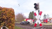 Ein Bahnübergang: Im Vordergrund das rot-weiße Andreaskreuz, im Hintergrund ein schwarzes Auto mit Totalschaden auf den Gleisen. © Screenshot 