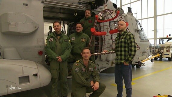 Ein junger Mann in zivil und mehrere Männer in Uniformen stehen neben einem Hubschrauber in einem Hangar. © Screenshot 