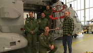 Ein junger Mann in zivil und mehrere Männer in Uniformen stehen neben einem Hubschrauber in einem Hangar. © Screenshot 