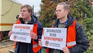 Zwei Klimaaktivisten der Gruppe "Letzte Generation": Sie stehen vor einem von ihnen orange angesprühten Weihnachtsbaum und halten Schilder in der Hand, auf denen steht: "Nächstenliebe = Klimaschutz!" und "Besinnlich in die Katastrophe?". © Screenshot 