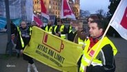 Kita-Beschäftigte demonstrieren in Wahnwesten und mit Fahnen auf einer Demo für höheren Lohn. © Screenshot 