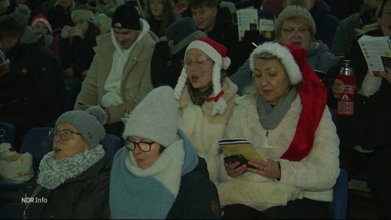 Mehrere Menschen sitzen im Stadion und singen, manche tragen rote Weihnachtsmützen. © Screenshot 