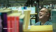 Hinter einem unscharf im Vordergrund erkennbaren Bücherregal steht Autor Till Raether und lacht herzlich. © Screenshot 