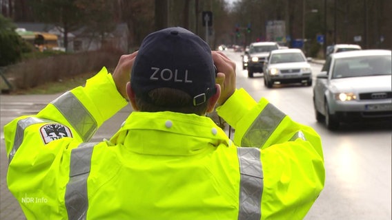 Ein Mann in neongelber Funktionsjacke trägt eine Cappie mit der Aufschrift "Zoll" und schaut durch ein Fernglas auf eine Straße auf der Autos vorbeifahren. © Screenshot 